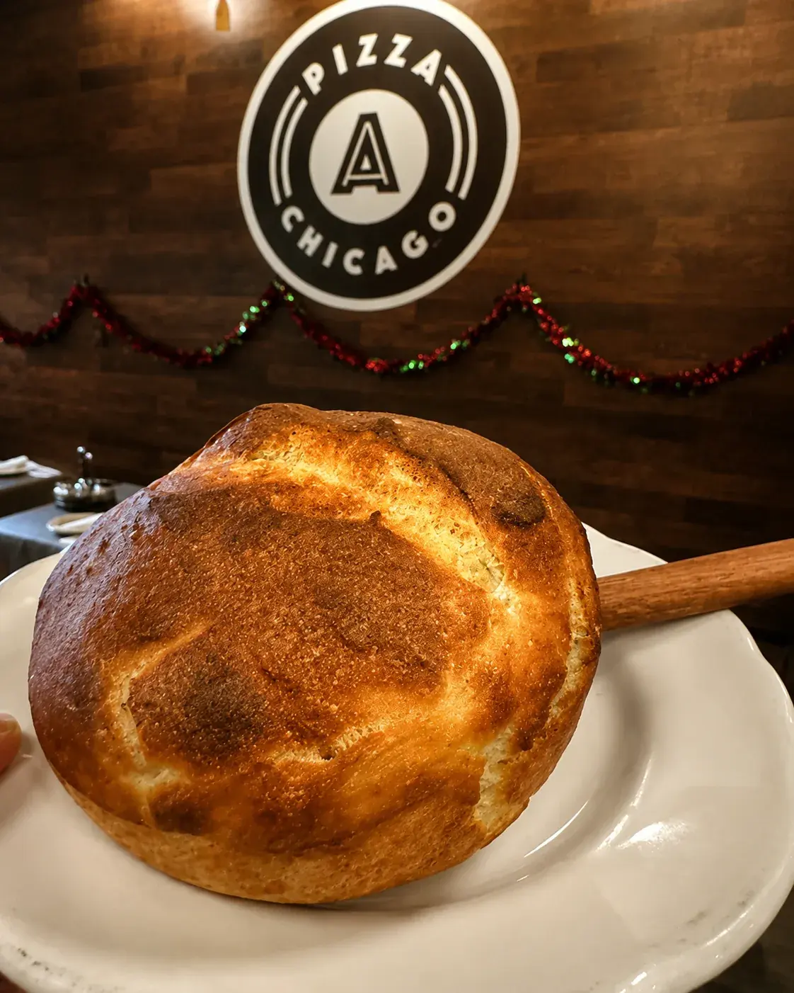 A round golden focaccia loaf resting on a wooden peel under the Anto Pizza Chicago logo
