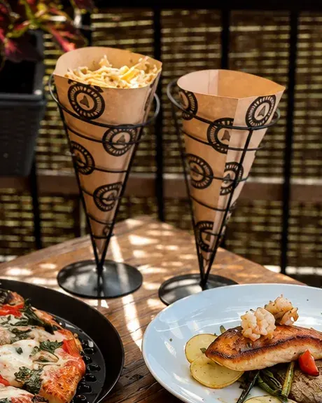 Two branded Anto paper cones on wire stands, one filled with fettuccine, on a sunlit patio table